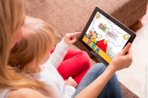 A young child sitting in her mother's lap playing a game on a tablet.